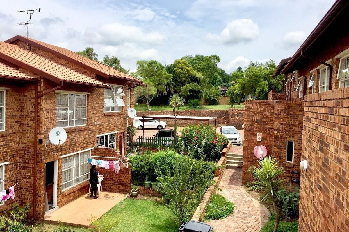 A view of the rear exterior of the apartment complex, featuring face brick buildings, a paved walkway, landscaped garden areas, and a communal carport.
