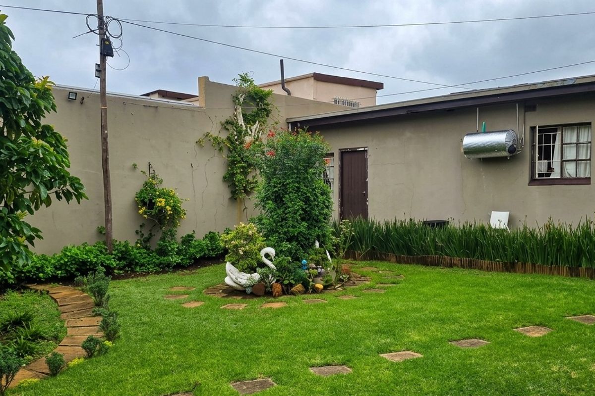 A rear garden area featuring a lawn, stone pathway, boundary wall, and an externally mounted solar geyser unit.