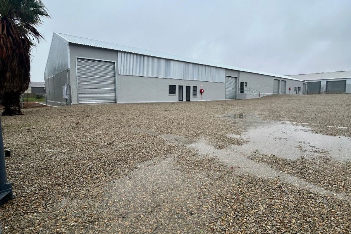 Rear view of a commercial warehouse facility featuring multiple roller shutter doors, metal cladding, and a large gravel yard area.