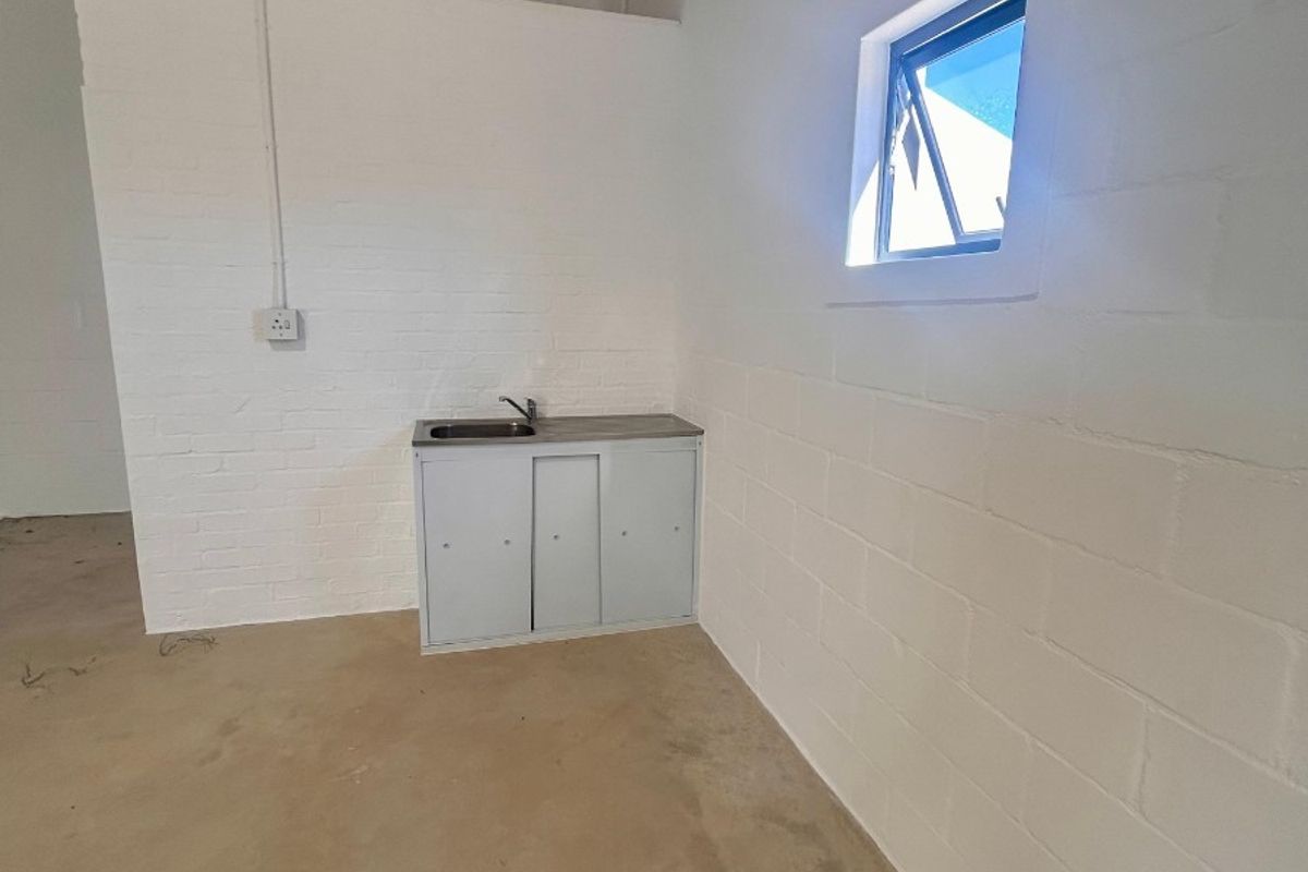 A minimalist retail kitchenette area featuring a compact sink unit, exposed brick walls, and high ceilings with exposed roof trusses.