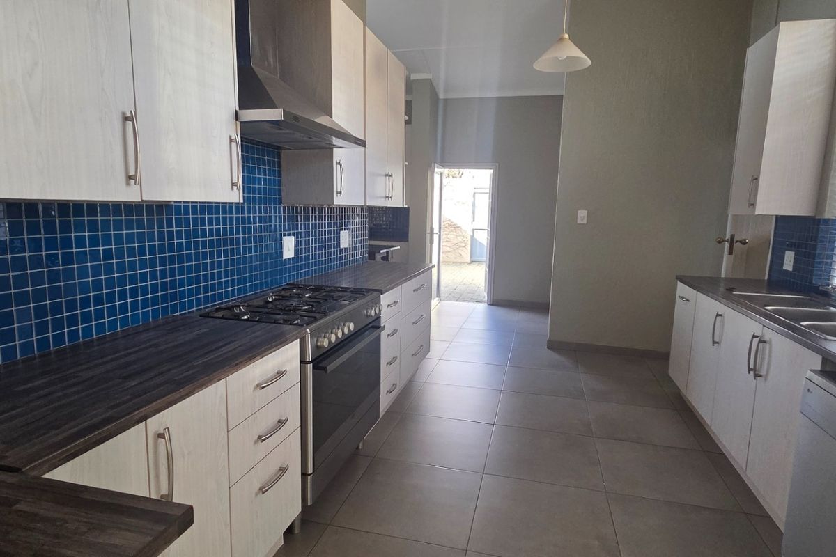 A functional kitchen featuring light-toned cabinetry, dark laminate countertops, a gas hob with extractor, and blue tiled backsplash, leading to an exterior doorway.