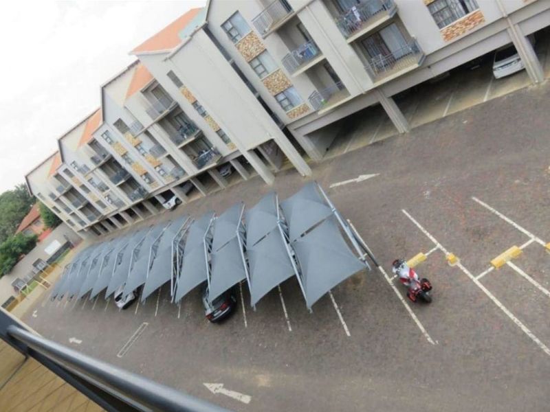 An elevated view of the apartment complex showing the building facade, balconies, and a row of covered parking bays in the paved parking area.