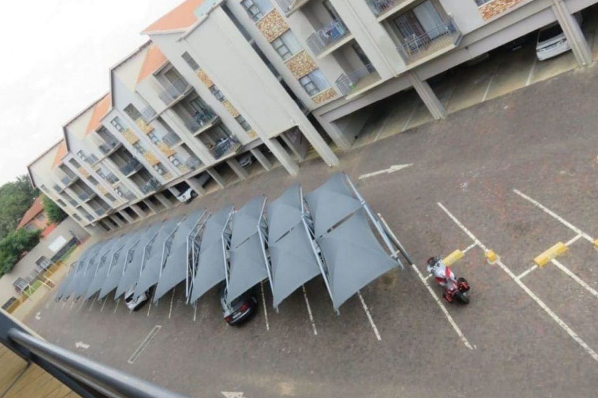 An elevated view of the apartment complex showing the building facade, balconies, and a row of covered parking bays in the paved parking area.