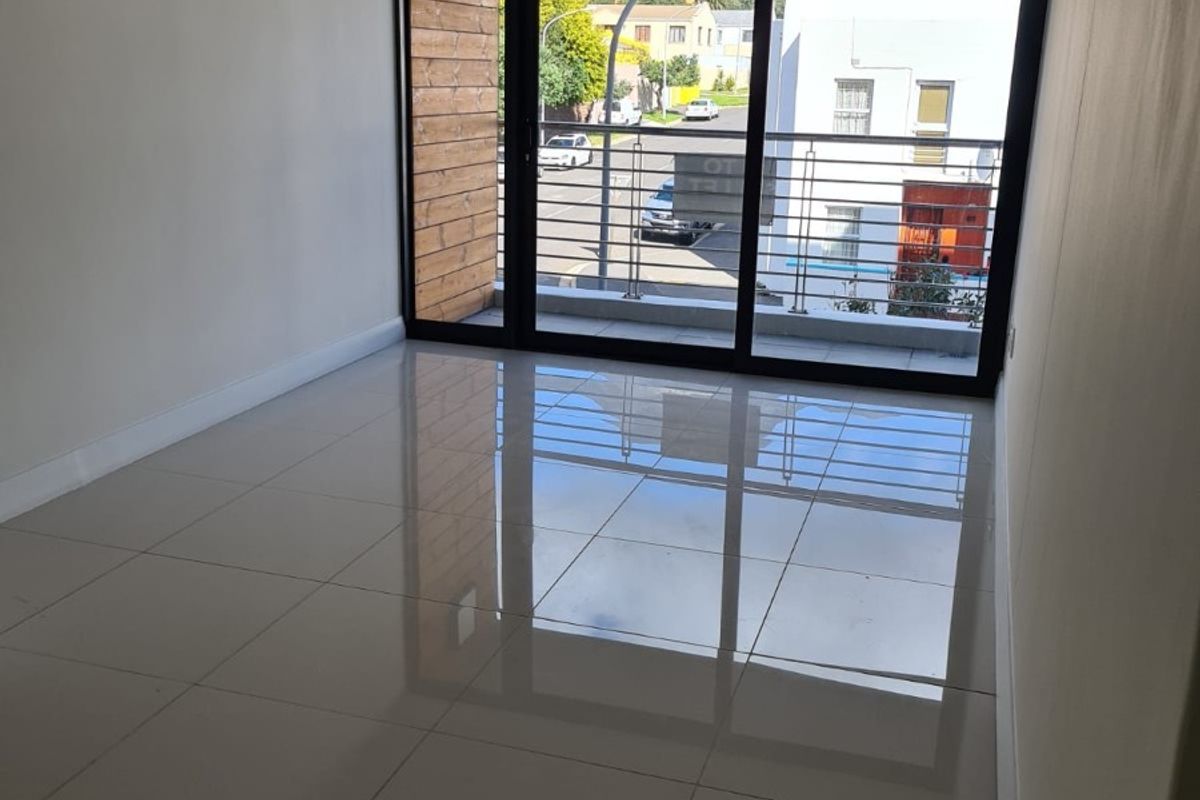 An empty, well-lit living area featuring polished tiled floors and a large sliding glass door leading to a balcony with a metal balustrade.