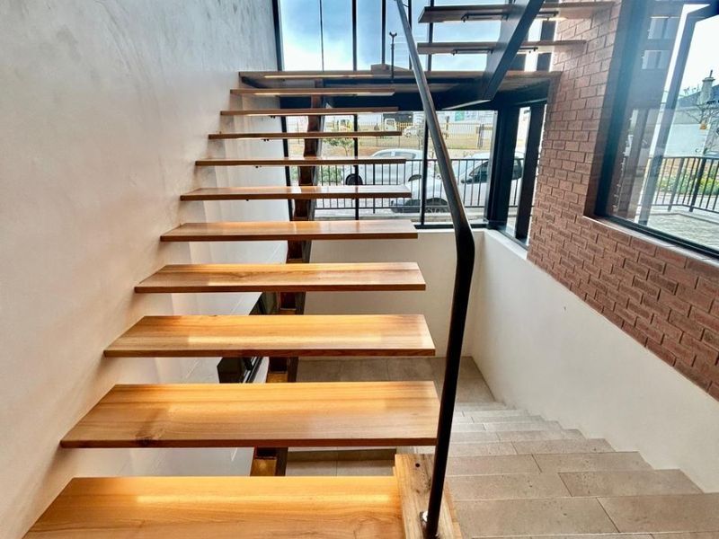 Modern architectural staircase featuring floating timber treads, steel handrails, and an exposed brick wall, illuminated by natural light through a large glass facade.