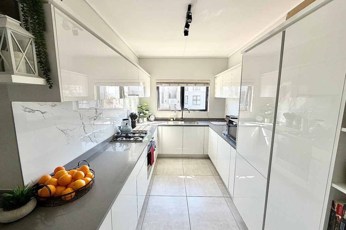 Modern galley-style kitchen featuring high-gloss white cabinetry, stone countertops, marble-look splashback, gas hob, and integrated appliances with neutral tiled flooring.