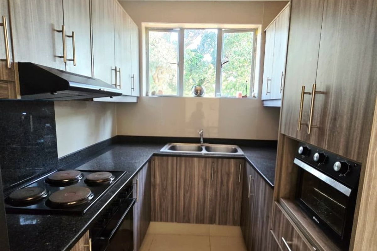 A modern, well-maintained galley kitchen featuring wood-grain cabinetry, dark countertops, built-in oven, electric hob, and a double bowl sink with natural light.