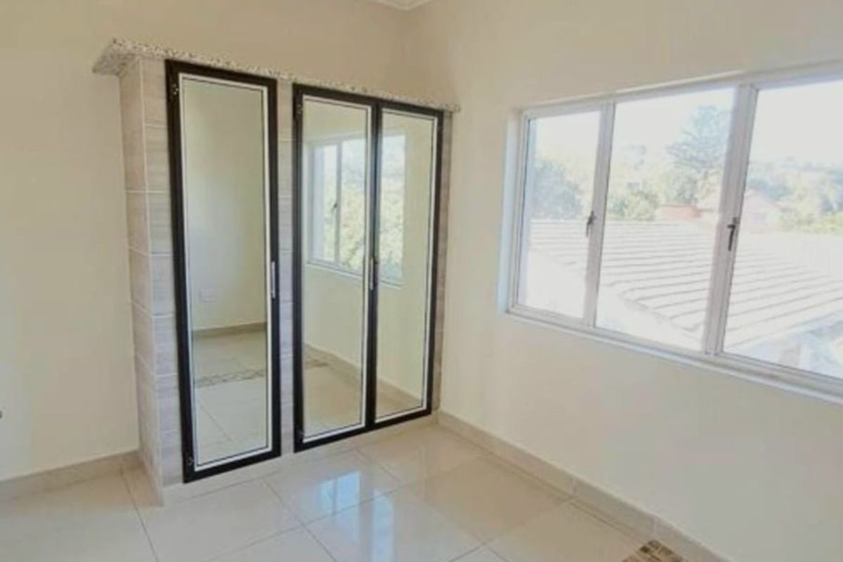 A bright, empty bedroom featuring built-in cupboards with mirrored doors, neutral tiled flooring, and large windows providing natural light.