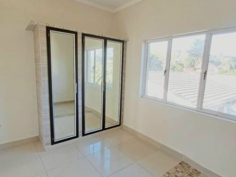 A bright, empty bedroom featuring built-in cupboards with mirrored doors, neutral tiled flooring, and large windows providing natural light.