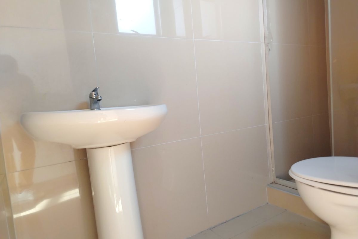 A clean, minimalist bathroom featuring a pedestal basin, chrome faucet, toilet, and a shower enclosure with neutral wall tiling.