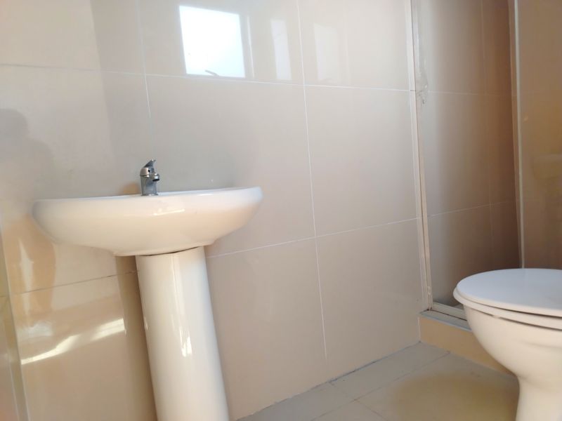 A clean, minimalist bathroom featuring a pedestal basin, chrome faucet, toilet, and a shower enclosure with neutral wall tiling.