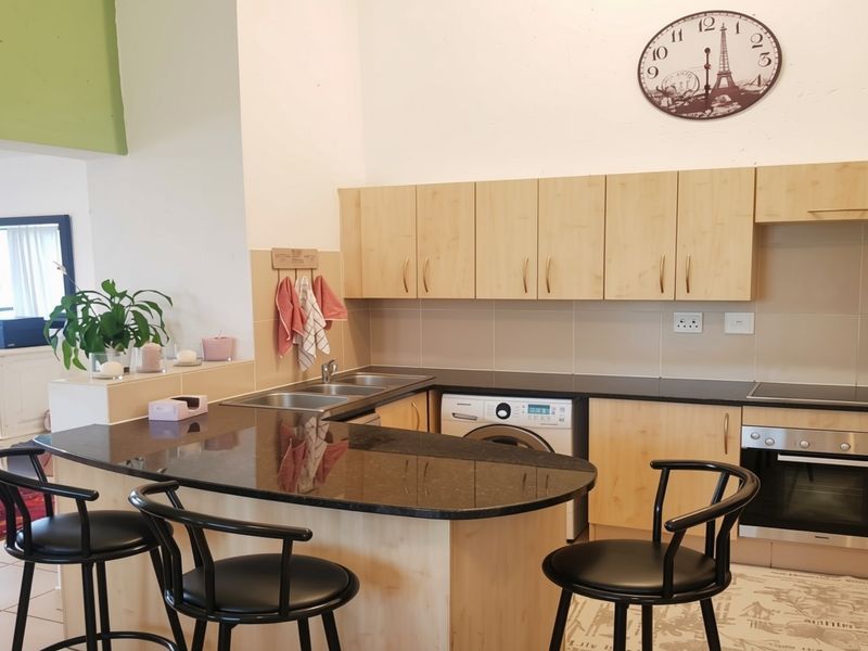 A functional kitchen featuring light wood cabinetry, dark granite countertops, a built-in oven, hob, and an integrated washing machine with a breakfast bar area.