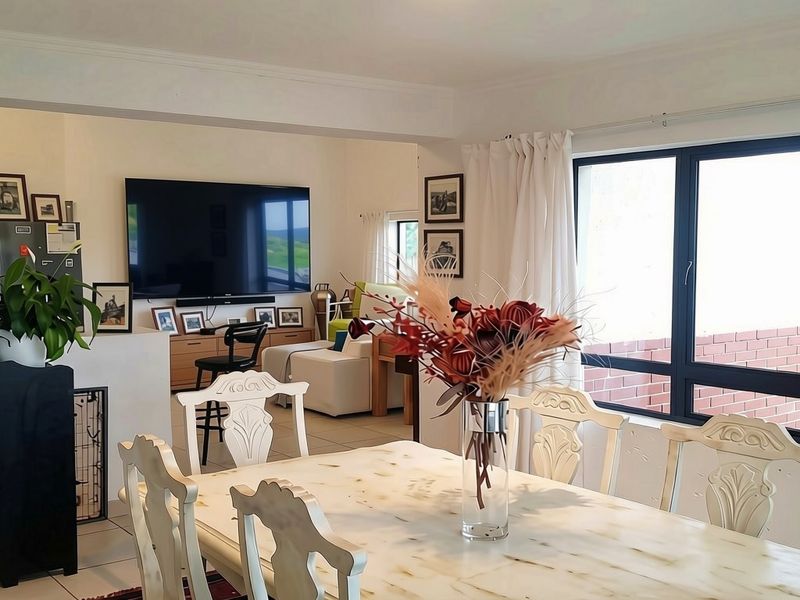 An open-plan living and dining area featuring tiled flooring, coved cornicing, and large aluminium-framed windows allowing for natural light.