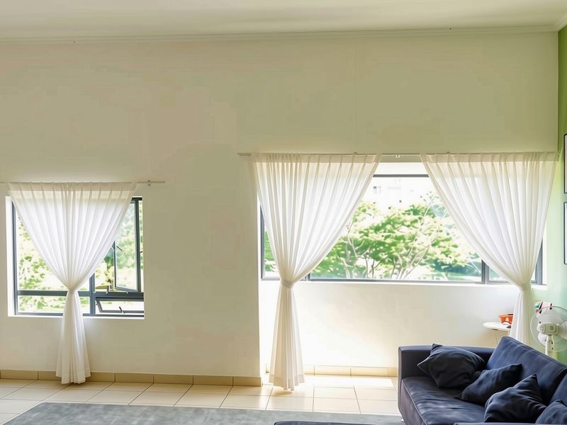 A bright living room featuring tiled flooring, large windows with sheer curtains, and a green accent wall, providing a clean and well-maintained living space.