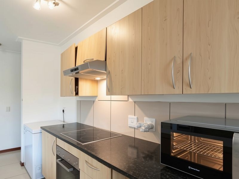 A functional kitchen featuring light wood-finish cabinetry, dark stone countertops, a built-in electric hob, oven, and extractor fan with neutral tiled splashbacks.