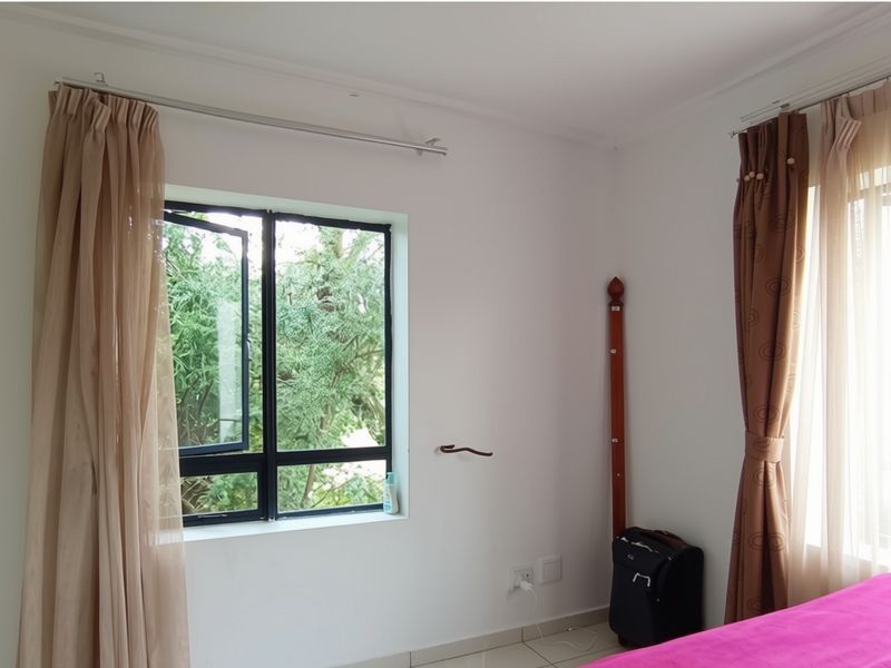 A bedroom featuring white walls, tiled flooring, and large windows with curtain rods, offering natural light and a view of the surrounding greenery.