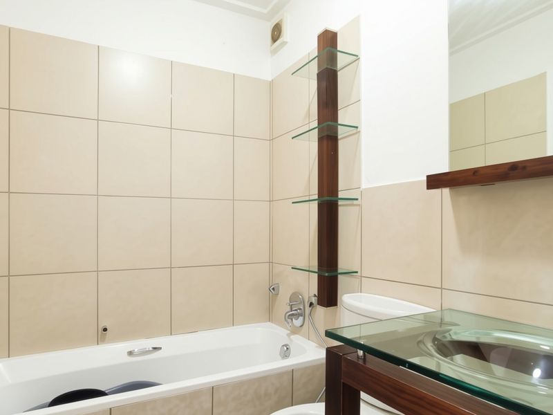 A modern bathroom featuring a bathtub, glass vanity basin, and integrated glass shelving with neutral wall tiling.