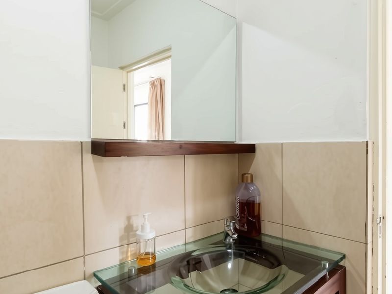 A bathroom vanity area featuring a glass vessel sink, chrome faucet, wall-mounted mirror, and a wooden shelf against neutral ceramic wall tiles.
