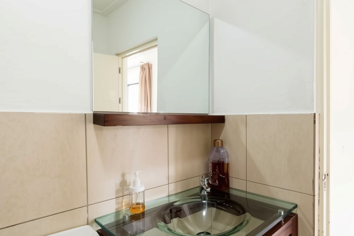 A bathroom vanity area featuring a glass vessel sink, chrome faucet, wall-mounted mirror, and a wooden shelf against neutral ceramic wall tiles.