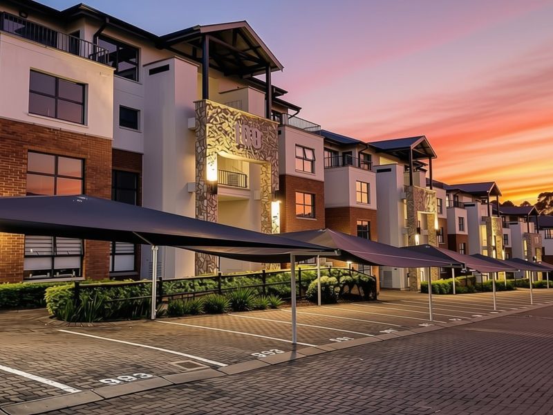 A well-maintained apartment complex exterior featuring brick and stone facade, paved parking bays with shade cloth carports, and private balconies at sunset.