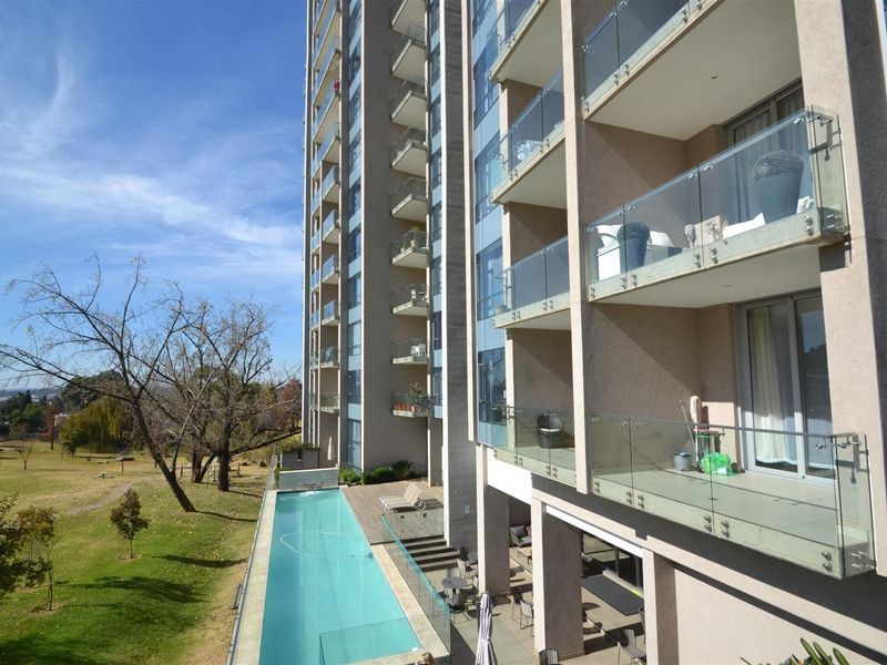 Exterior view of a modern apartment building featuring multiple balconies with glass balustrades, overlooking a swimming pool and landscaped grounds.