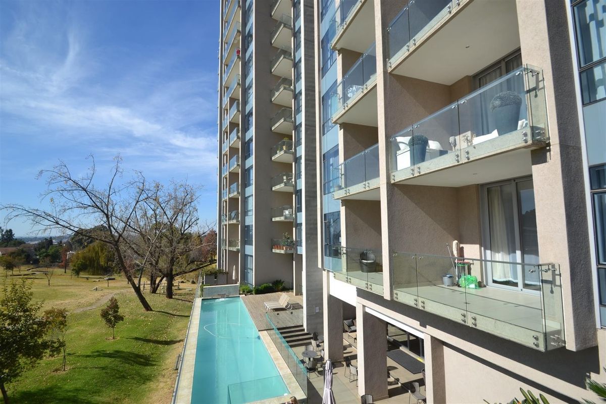 Exterior view of a modern apartment building featuring multiple balconies with glass balustrades, overlooking a swimming pool and landscaped grounds.