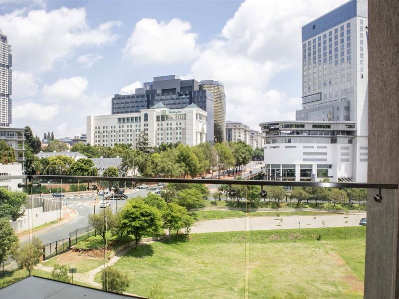 An elevated urban view overlooking a green space and surrounding city buildings, featuring a modern glass balustrade in the foreground.