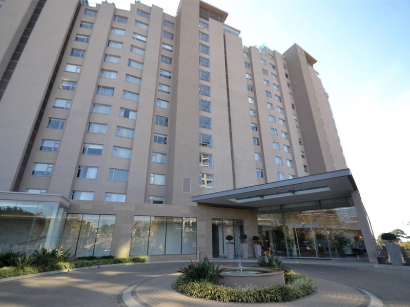 Front exterior view of a high-rise apartment building featuring a paved driveway, central fountain, and a modern glass-fronted entrance canopy.