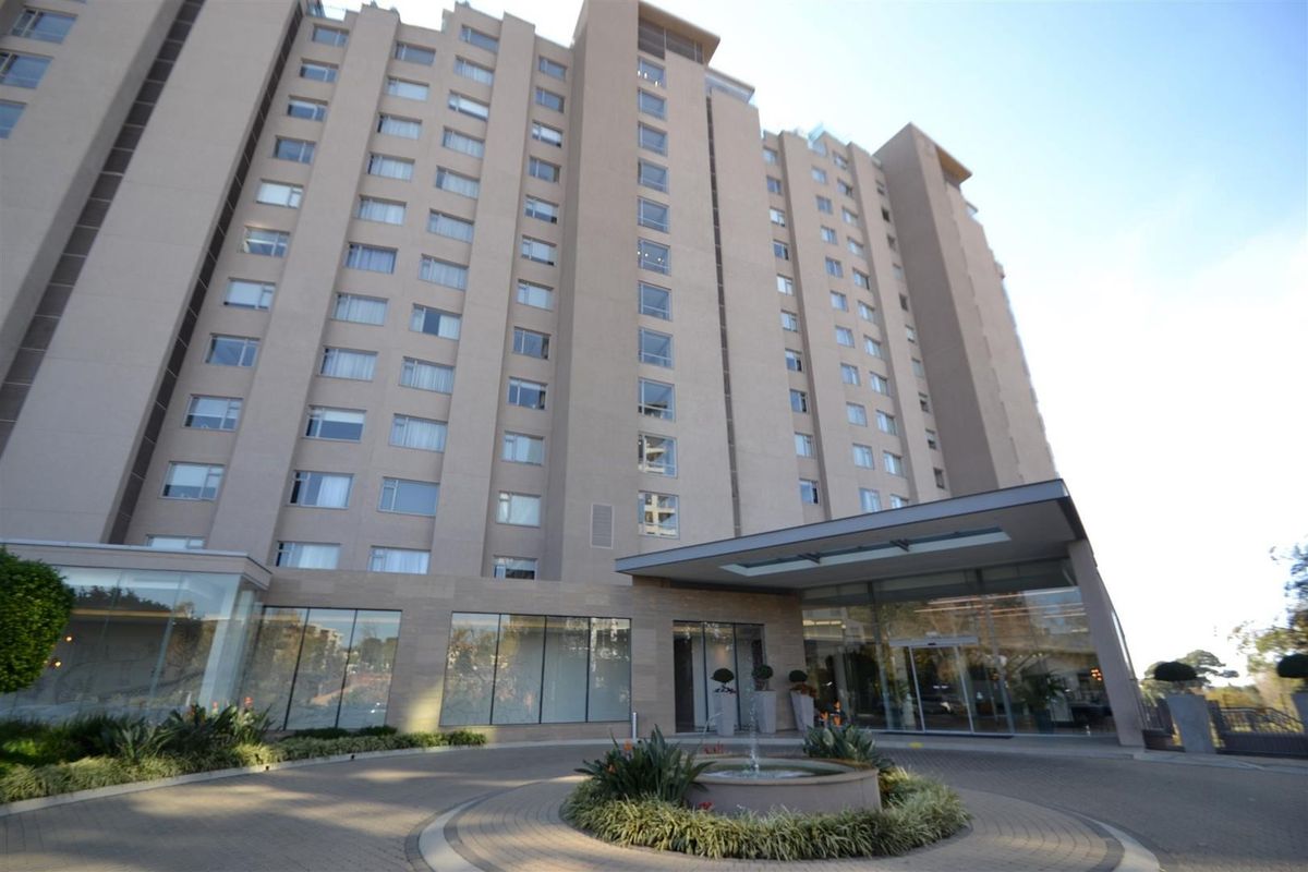 Front exterior view of a high-rise apartment building featuring a paved driveway, central fountain, and a modern glass-fronted entrance canopy.