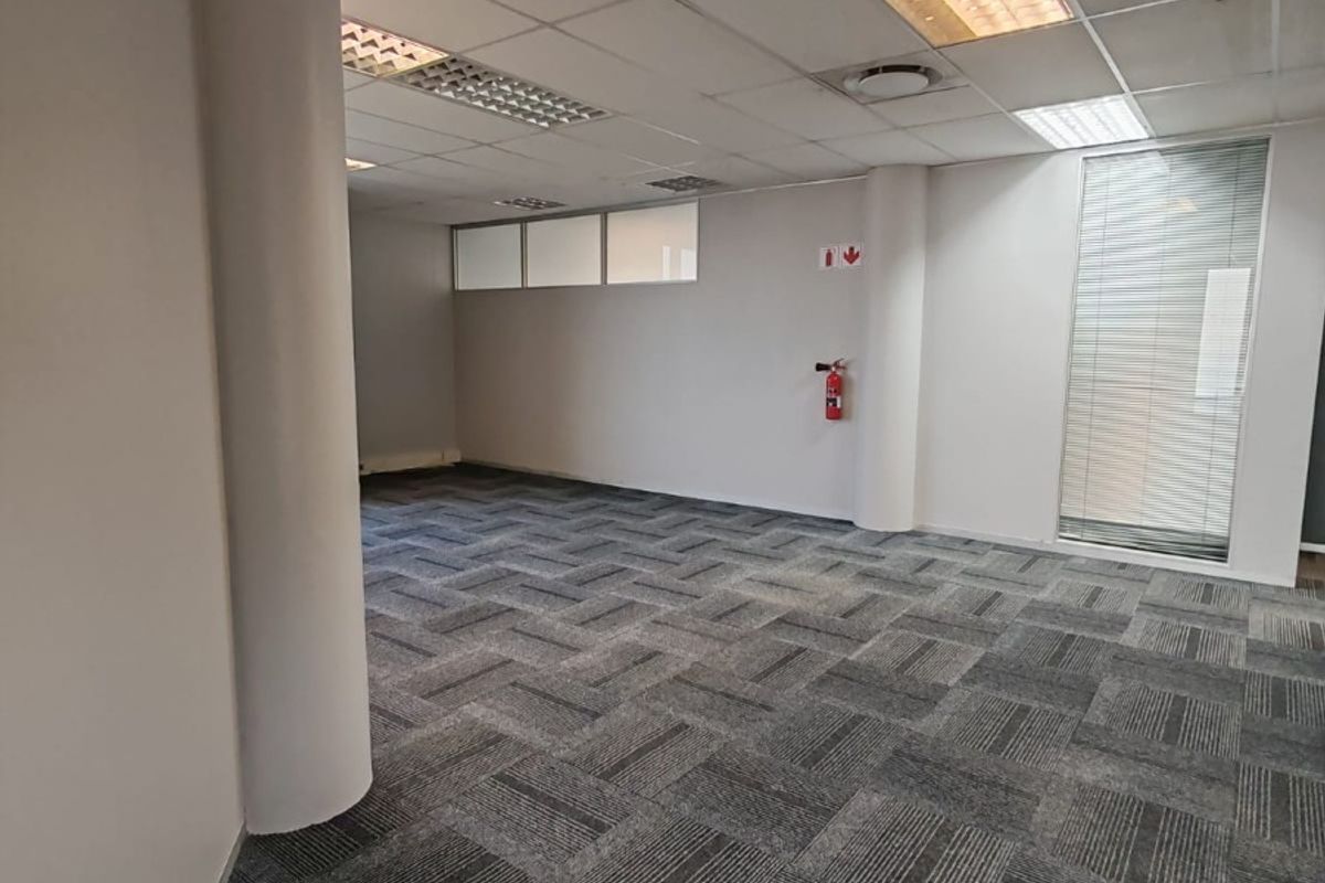 A vacant, open-plan office space featuring carpet tiles, a suspended ceiling with recessed lighting, and a structural column.