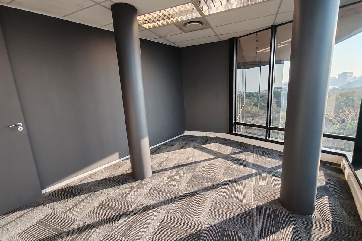 A bright, empty office space featuring modern carpet tiles, structural columns, and large windows providing ample natural light and exterior views.