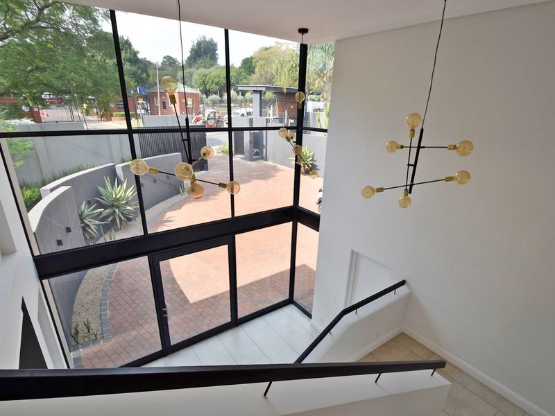 A bright, double-volume entrance hall featuring floor-to-ceiling windows, modern pendant lighting, and a staircase overlooking the paved front courtyard.