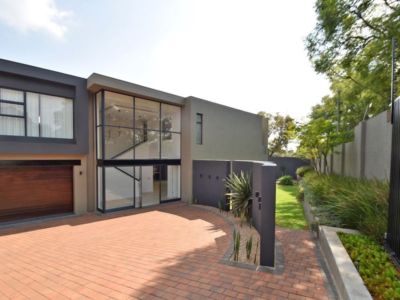 Modern double-storey residence featuring a striking glass facade, paved driveway, timber garage door, and well-maintained landscaped garden with secure perimeter fencing.