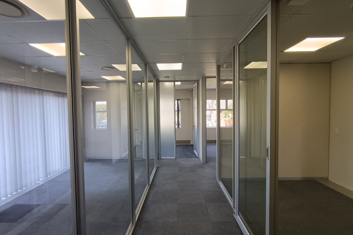 A bright, modern office hallway featuring glass partition walls, suspended ceilings with recessed lighting, and clean carpet flooring.