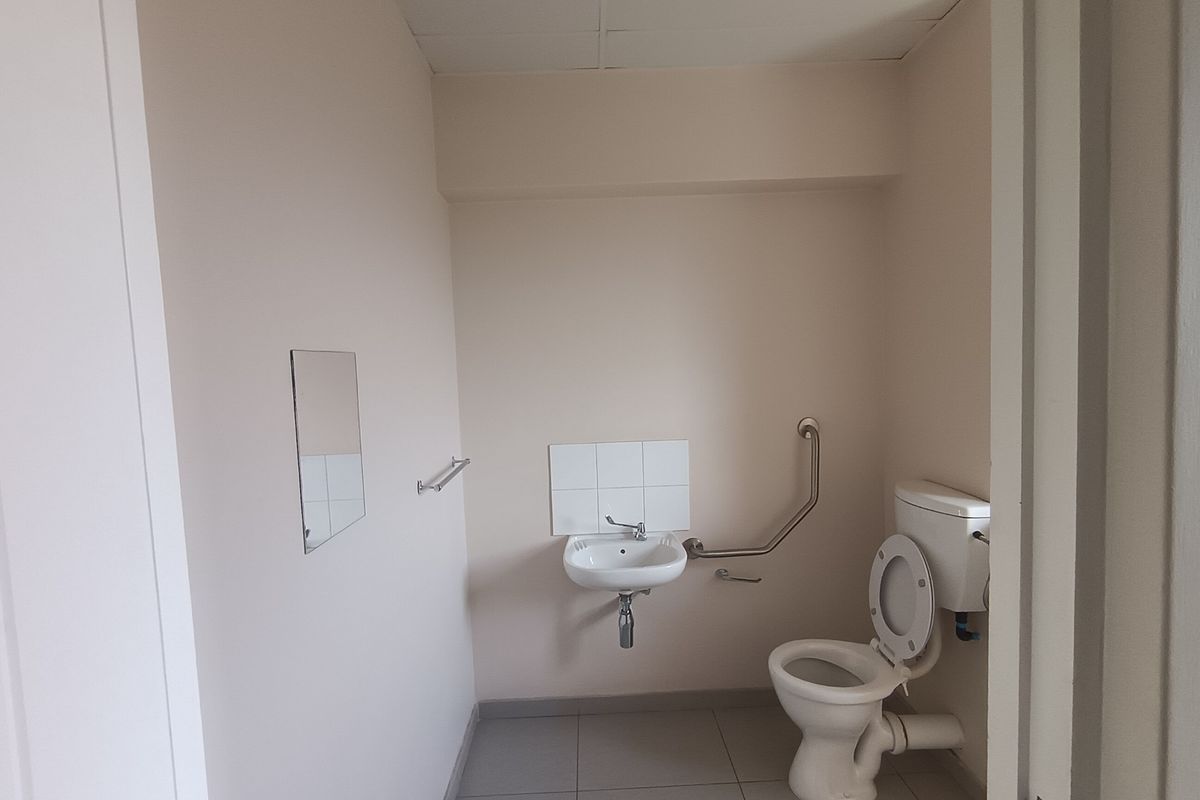 A functional, accessible office bathroom featuring a wall-mounted basin, toilet, and safety grab rail with neutral wall finishes and tiled flooring.