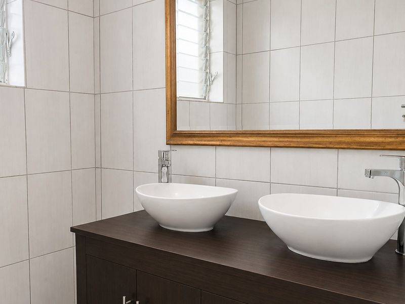 A modern bathroom featuring a dark wood double vanity with white vessel sinks, chrome fixtures, and full-height wall tiling.