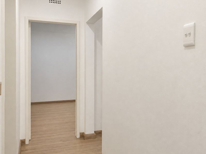 A clean, neutral hallway featuring light-toned laminate flooring, white walls, and simple door frames leading into an adjacent room.