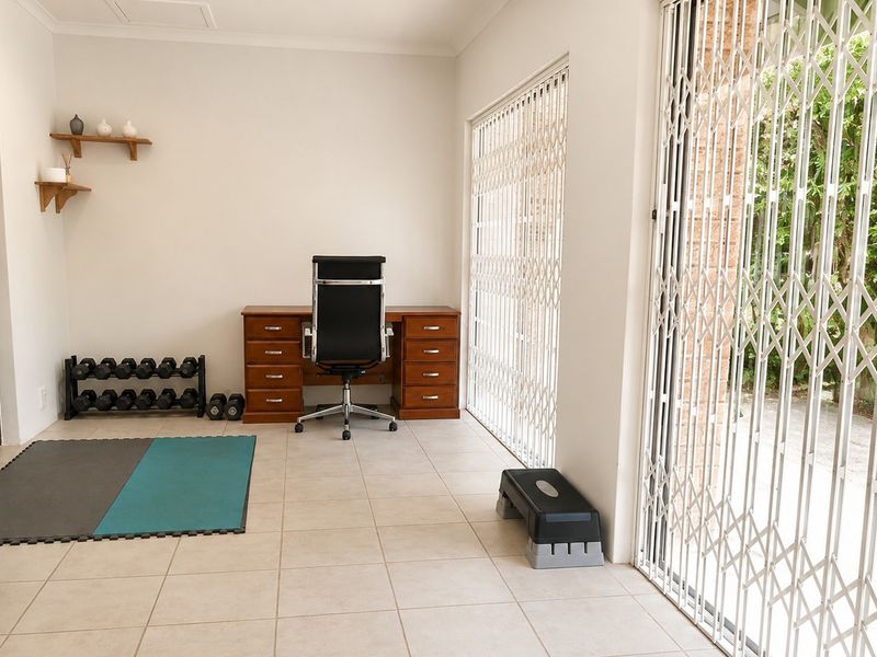 A bright home office space featuring tiled flooring, floating wall shelves, and large sliding glass doors secured with white metal security gates.