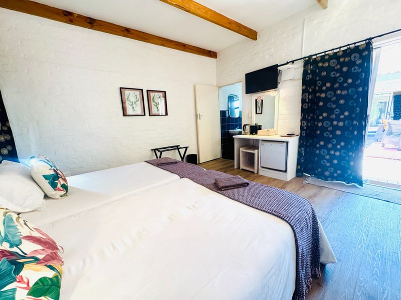 A bright bedroom featuring exposed timber ceiling beams, painted brick walls, laminate flooring, and direct access to an en-suite bathroom and outdoor area.