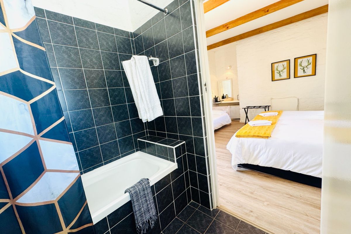 A bathroom featuring dark tiled walls and a built-in bathtub, with an open transition into a bedroom area with exposed timber ceiling beams.