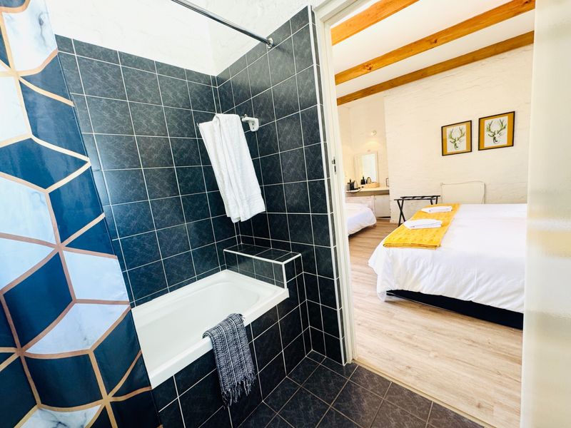 A bathroom featuring dark tiled walls and a built-in bathtub, with an open transition into a bedroom area with exposed timber ceiling beams.