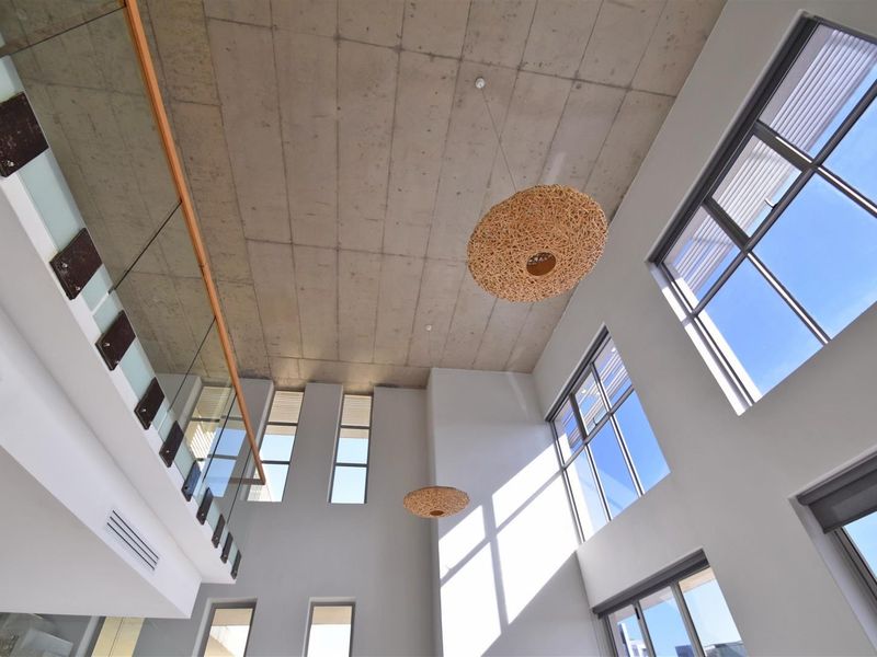 Spacious double-volume living area featuring an exposed concrete ceiling, large windows for natural light, and a modern glass balustrade overlooking the space.
