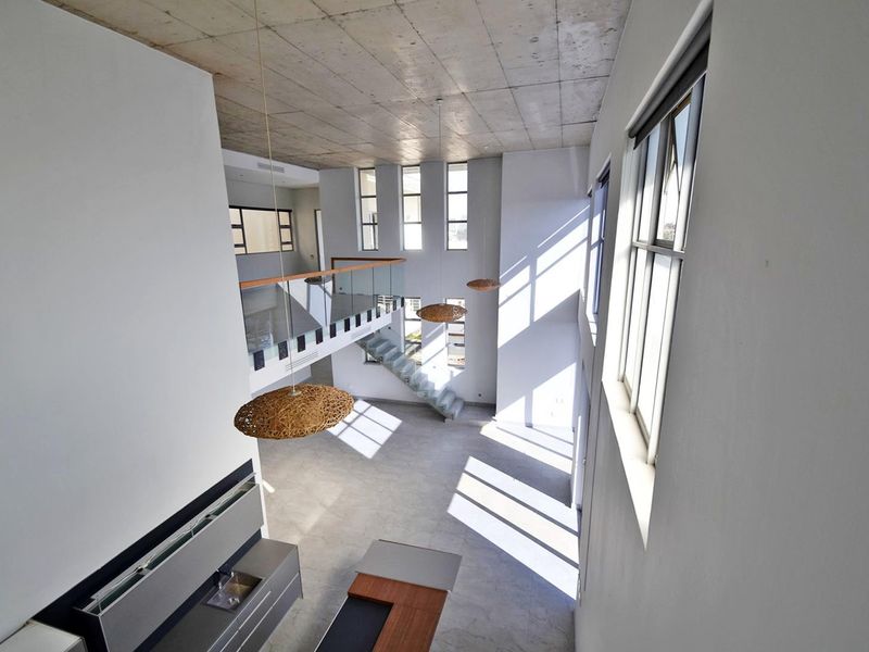 Spacious open-plan living area featuring high ceilings, exposed concrete finishes, a modern staircase with glass balustrade, and abundant natural light.