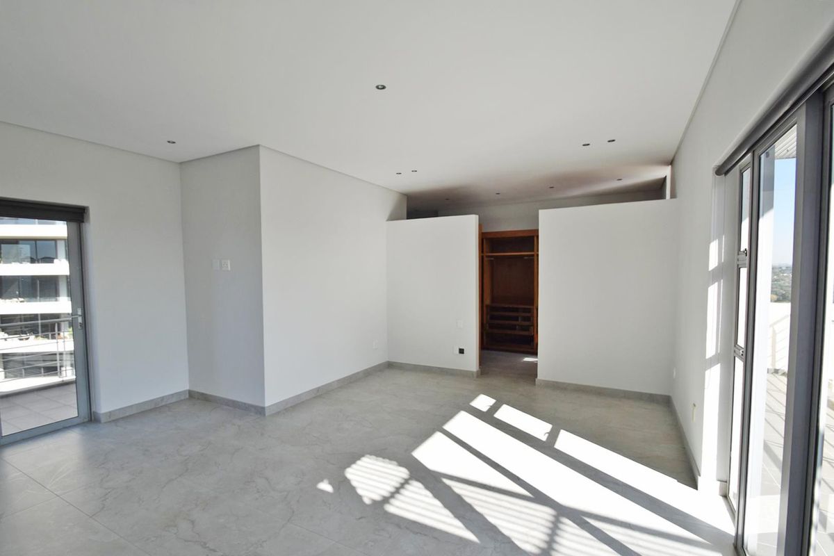 A bright, modern bedroom featuring neutral tiled flooring, recessed lighting, and built-in wardrobe space, with direct access to a balcony through sliding glass doors.