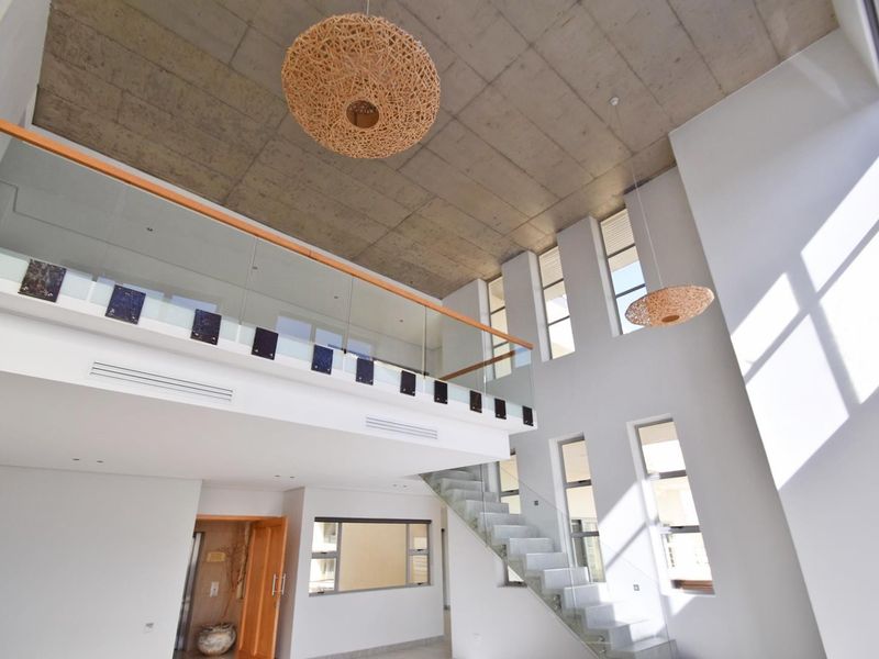 Spacious double-volume living area featuring an exposed concrete ceiling, modern glass staircase, and large vertical windows providing abundant natural light.