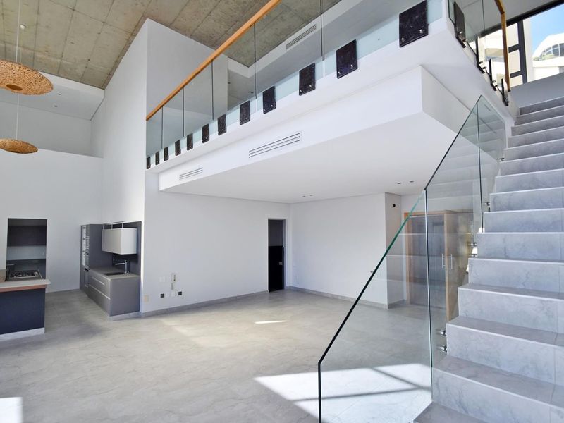 Spacious open-plan living area featuring a modern kitchen, double-volume exposed concrete ceiling, and a striking glass-railed staircase leading to a mezzanine level.