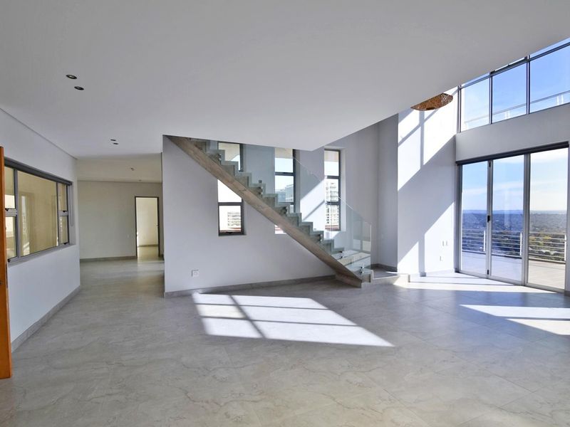 Spacious double-volume living area featuring modern tiled flooring, a contemporary staircase with glass balustrade, and large windows providing ample natural light and balcony access.