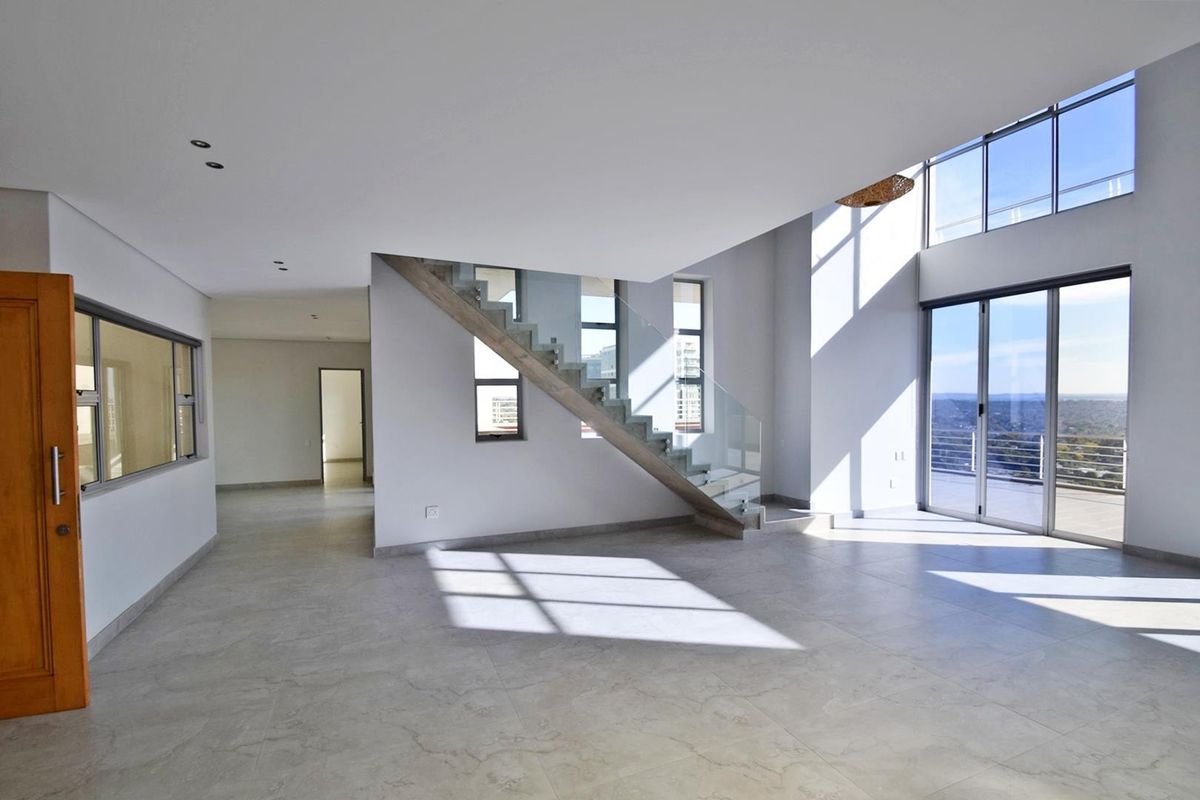 Spacious double-volume living area featuring modern tiled flooring, a contemporary staircase with glass balustrade, and large windows providing ample natural light and balcony access.