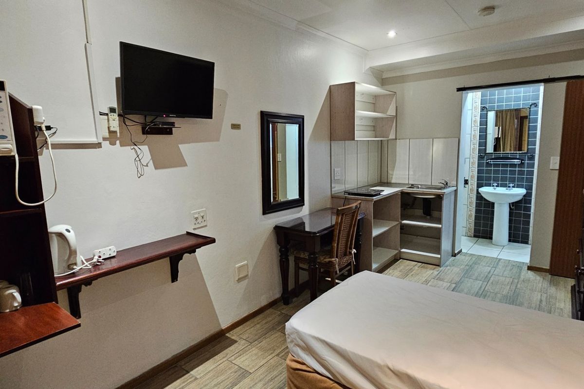 A compact bedroom featuring a kitchenette area with sink and an adjoining en-suite bathroom with a pedestal basin and tiled finishes.