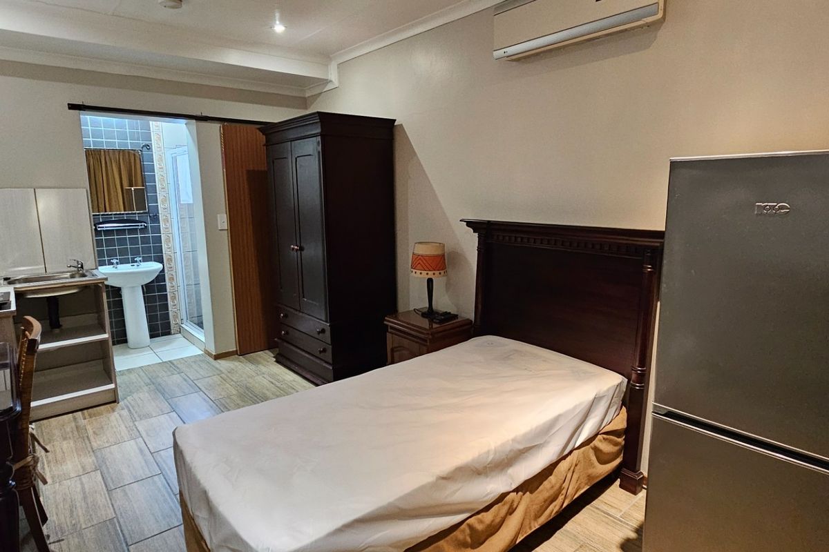 A compact bedroom featuring an integrated kitchenette, tiled flooring, split-system air conditioning, and an en-suite bathroom with a shower and pedestal basin.